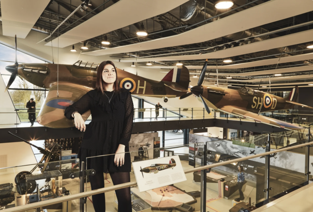 Giant woman stands resting her arm on a Spitfire in the Battle of Britain museum
