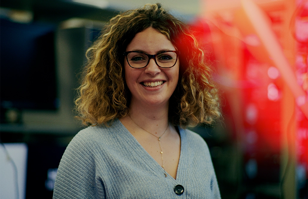 Hitachi Woman glasses grey top & curly hair smiling into camera